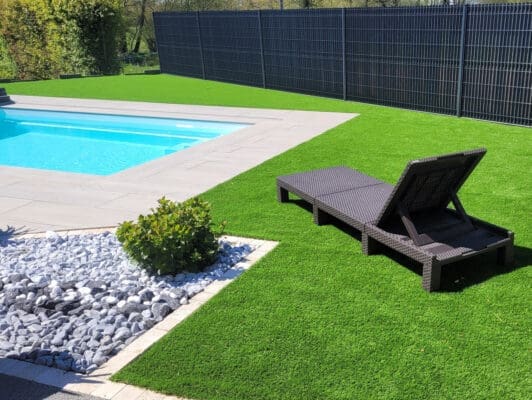 Aménagement extérieur avec galets décoratifs gris et blancs, gazon synthétique et terrasse en pierre autour d’une piscine, réalisé avec les produits Ozaé.